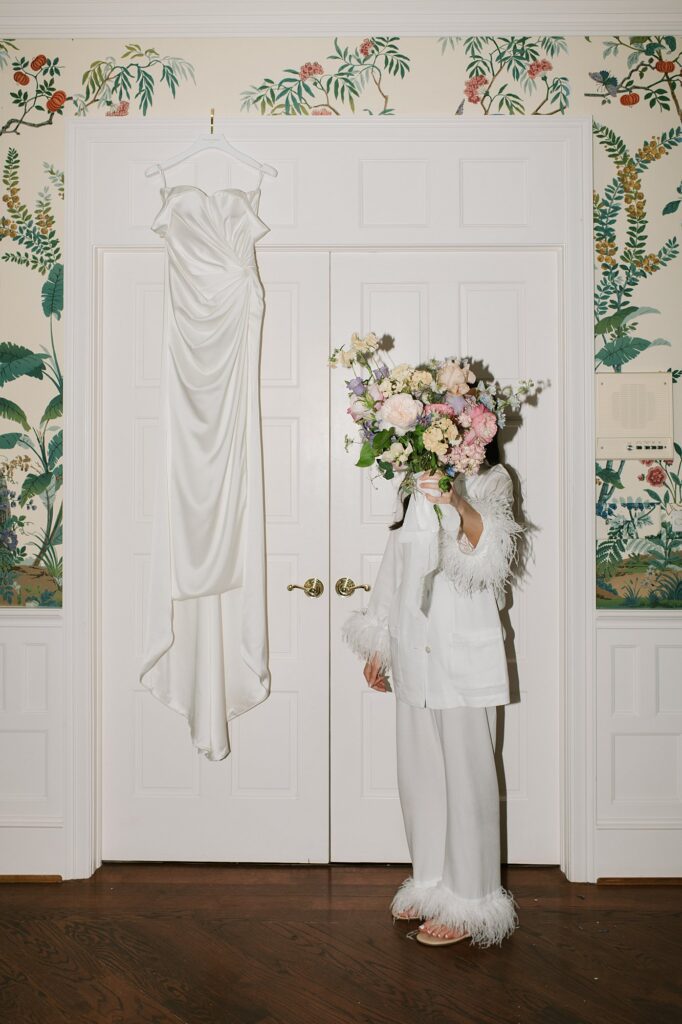 a bride holding her bouquet in front of her face next to her dress hanging from a door before her Greystone Castle wedding